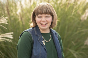 Young woman with big smile, shoulder length brown hair, denim vest and green shirt stands in front of sunlit tall grasses