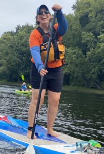 A woman wearing an orange t-shirt and a yellow life jacket smiles while paddleboarding