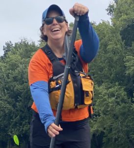 A woman in an orange shirt, sunglasses, and baseball cap smiles while taking a paddle boarding stroke