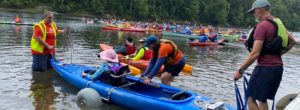 A woman helps to launch a blue tandem kayak as a larger flotilla is waiting in the background