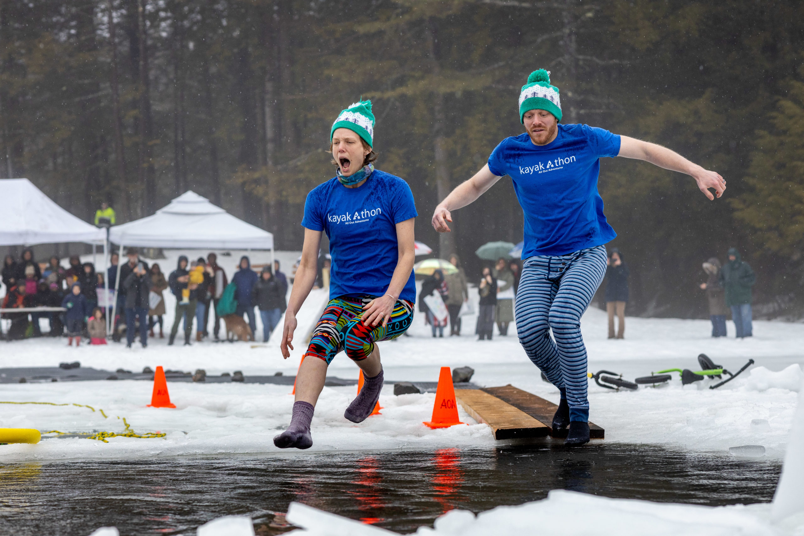 2 people wearing blue "Kayak-a-thon" t-shirts and green and white hats jump into the plunge hole