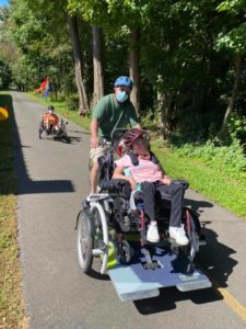 Image is of a woman in a wheelchair tandem bike while a man pedals behind her