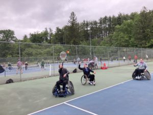 3 people in wheelchairs are behind the baseline on a tennis court as one serves