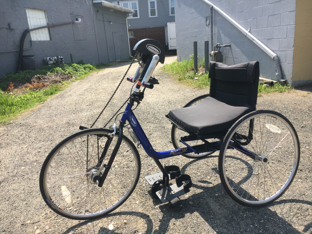 Blue handcycle with the seat higher up off the ground and upright seat position. There are footrests, hand cranks to pedal, and one wheel in the front and two wheels in the back. 