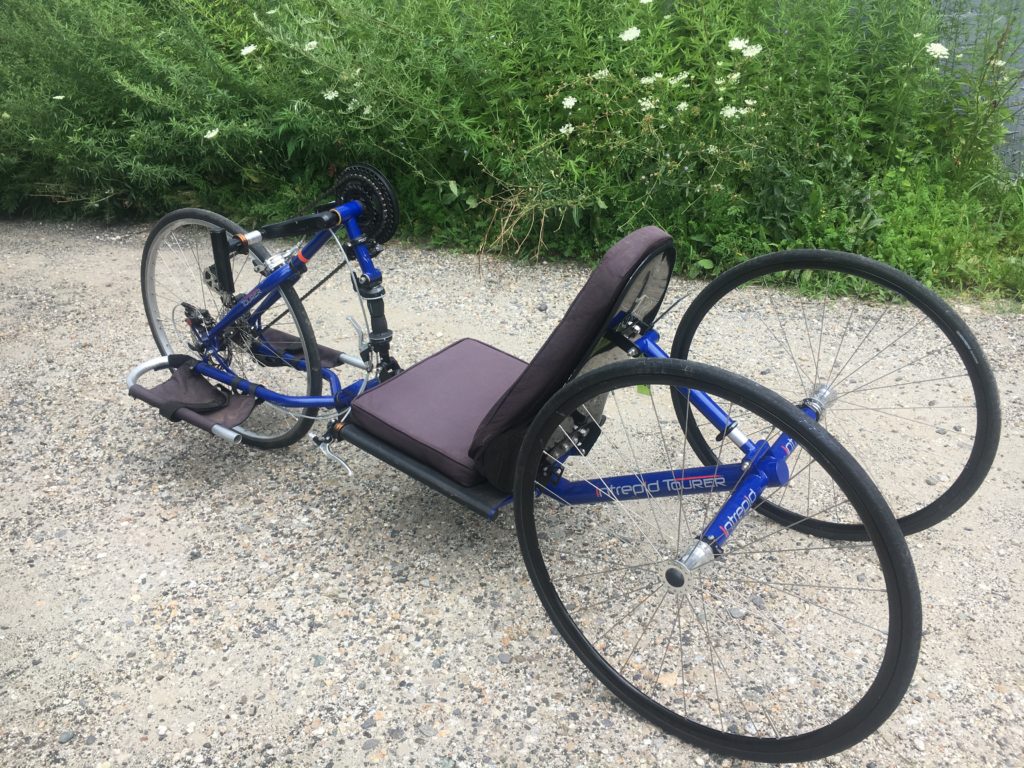 A blue recumbent handcycle. The seat is a few inches off the ground and there is one wheel in the front and two wheels in the back. There are foot rests and hand levers in the front to propel the cycle forward. 