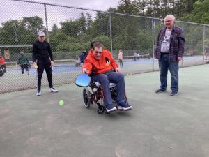 A person using a wheelchair swings a pickleball paddle at a ball while others look on near him