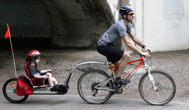 A person pedaling an upright bicycle with the trailer attached to the bike seat post. A child is sitting in the trailer and pedaling. 