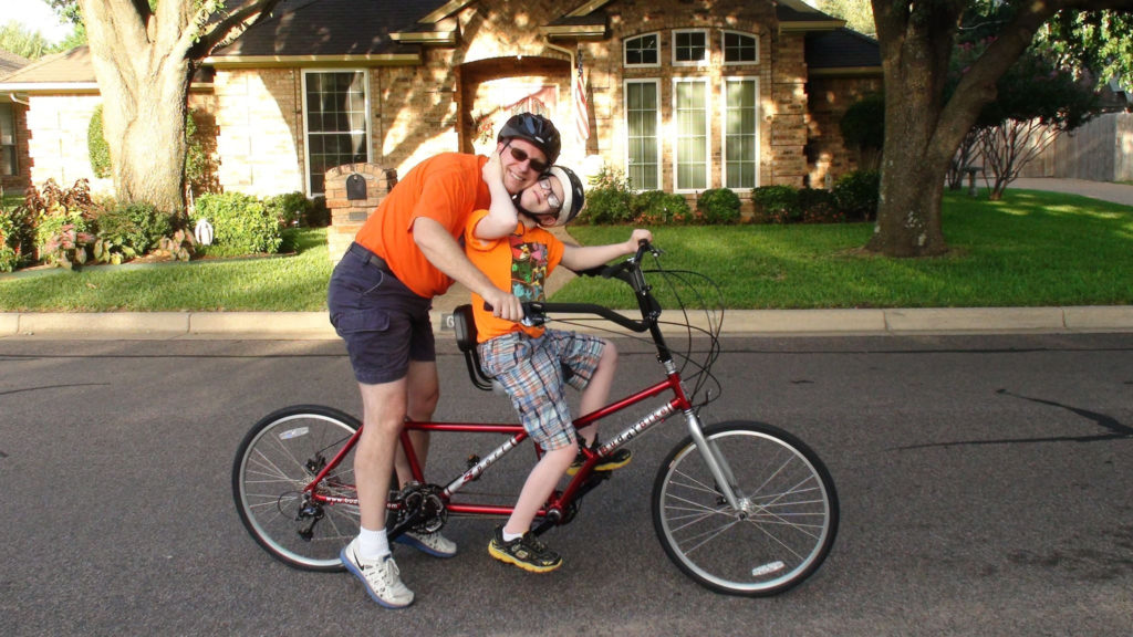 Two riders on the Buddy Bike, with the rider in back taller than the rider in the front. 