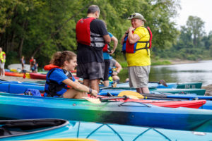A young girl sitting in a kayak on land with 2 adults standing and talking next to her
