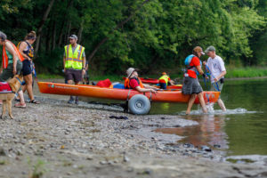 A kayak with paddler being wheeled into the water on large "chariot" wheels