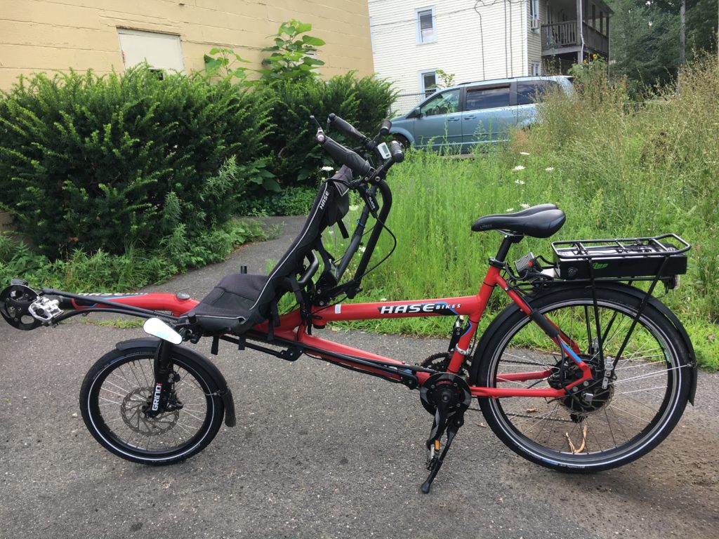 A red tandem bicycle. The front seat is recumbent and has a shorter wheel, the back seat is an upright bicycle seat and has a larger wheel. There is also an electric assist battery on a rear rack over the back wheel. 