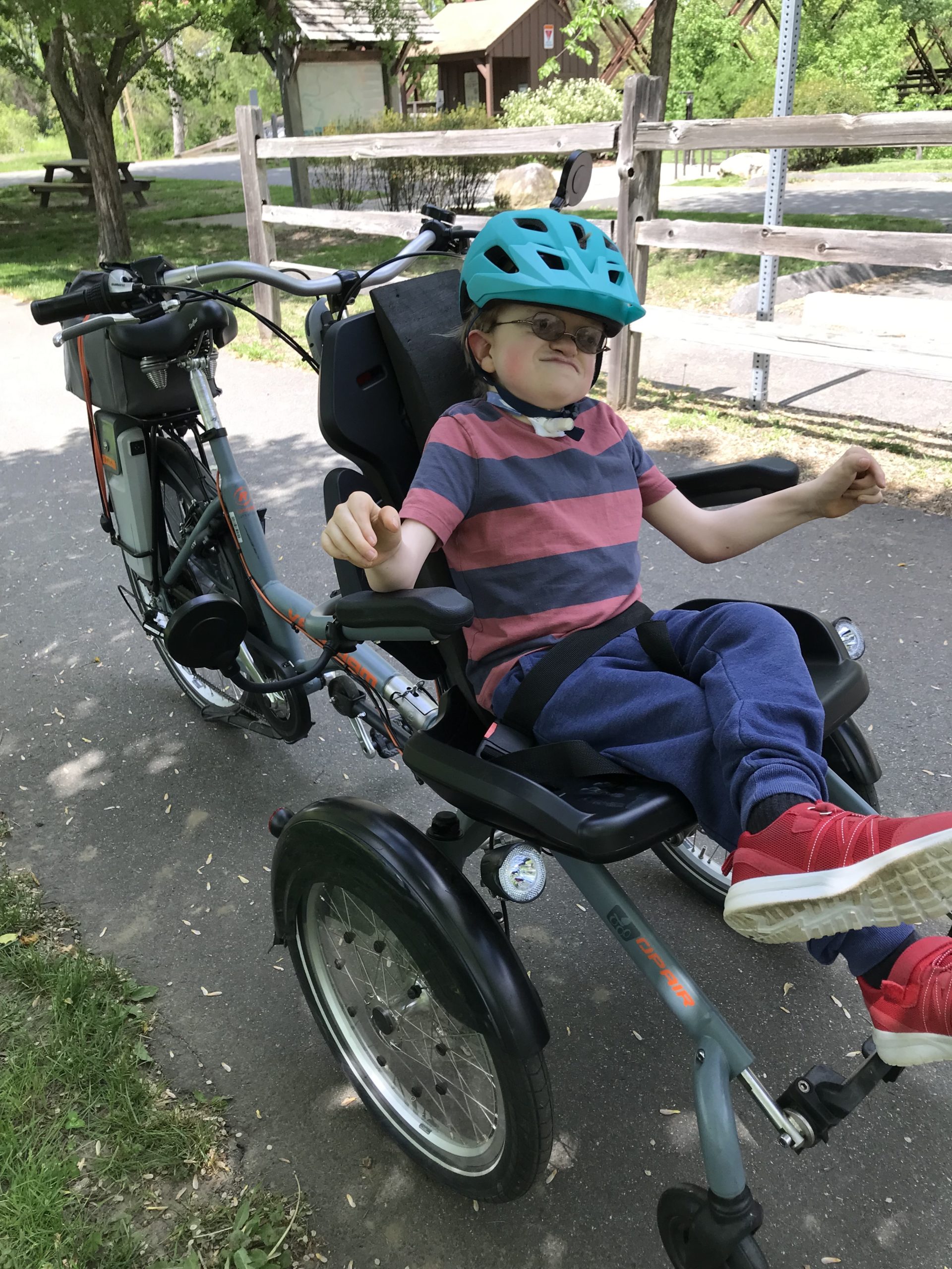 A person sitting in a wheelchair. Attached to the back of a chair is a bike seat, wheel, handlebars, and pedals.