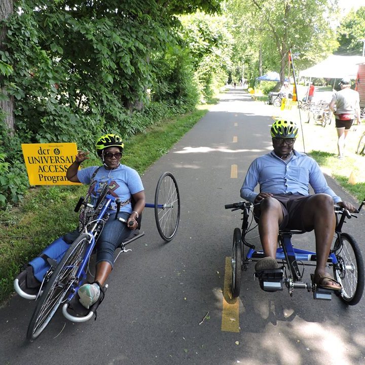 2 people, smiling, one on a handcycle and one on a recumbent trike