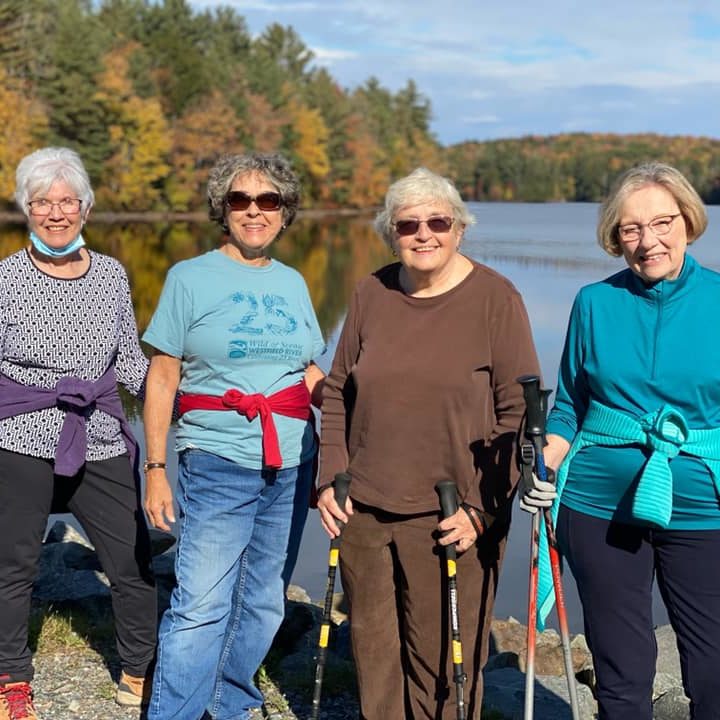 4 women stand in front of a calm pond, smiling, with fall foliage behind them